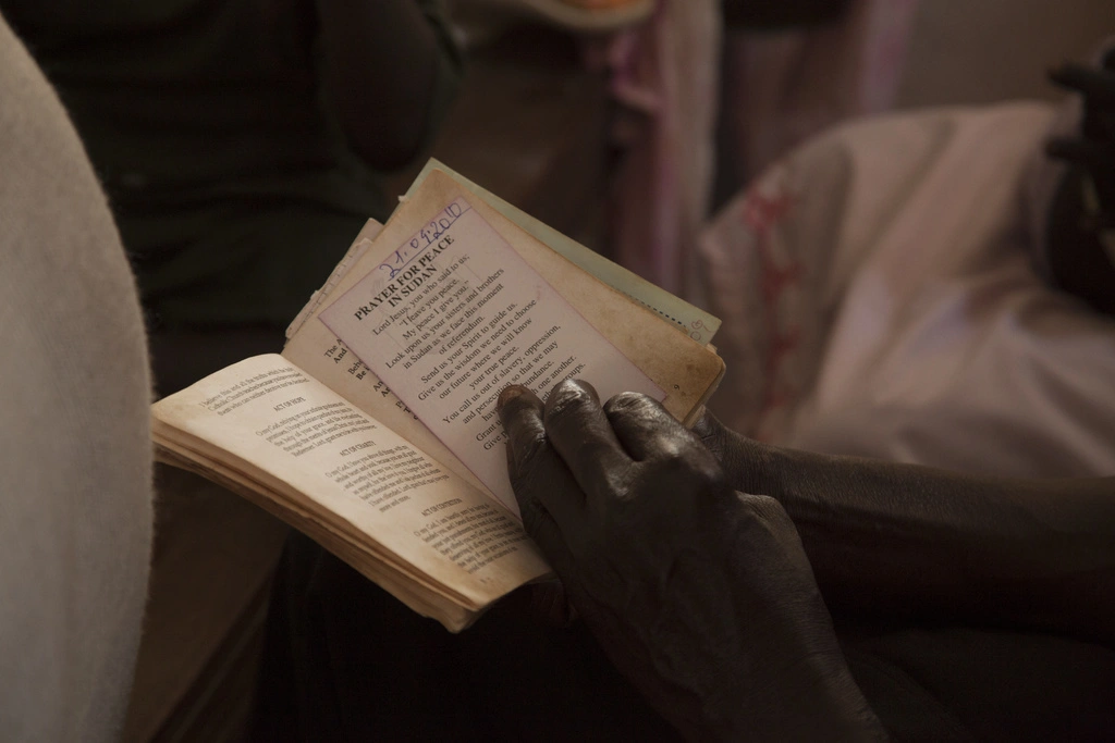 A person opening a book with a slip of paper inside of it titled 'Prayer for Peace in Sudan' with the written prayer under the title