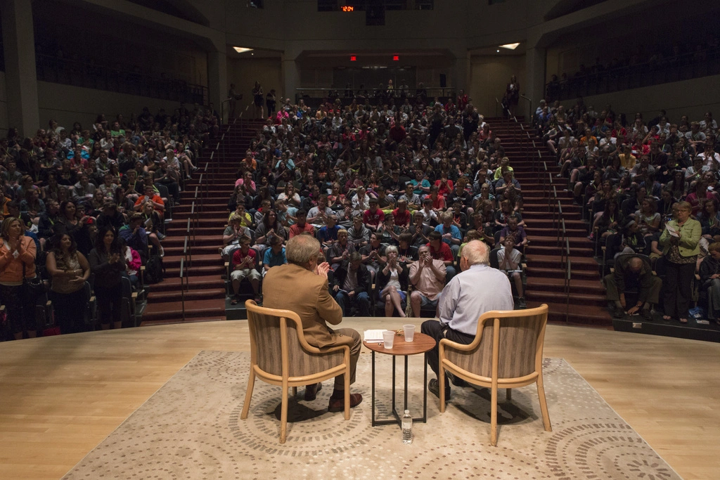 A Holocaust survivor and interviewer sit in chairs in one of the Museum's auditoriums, while an audience applauds in the background